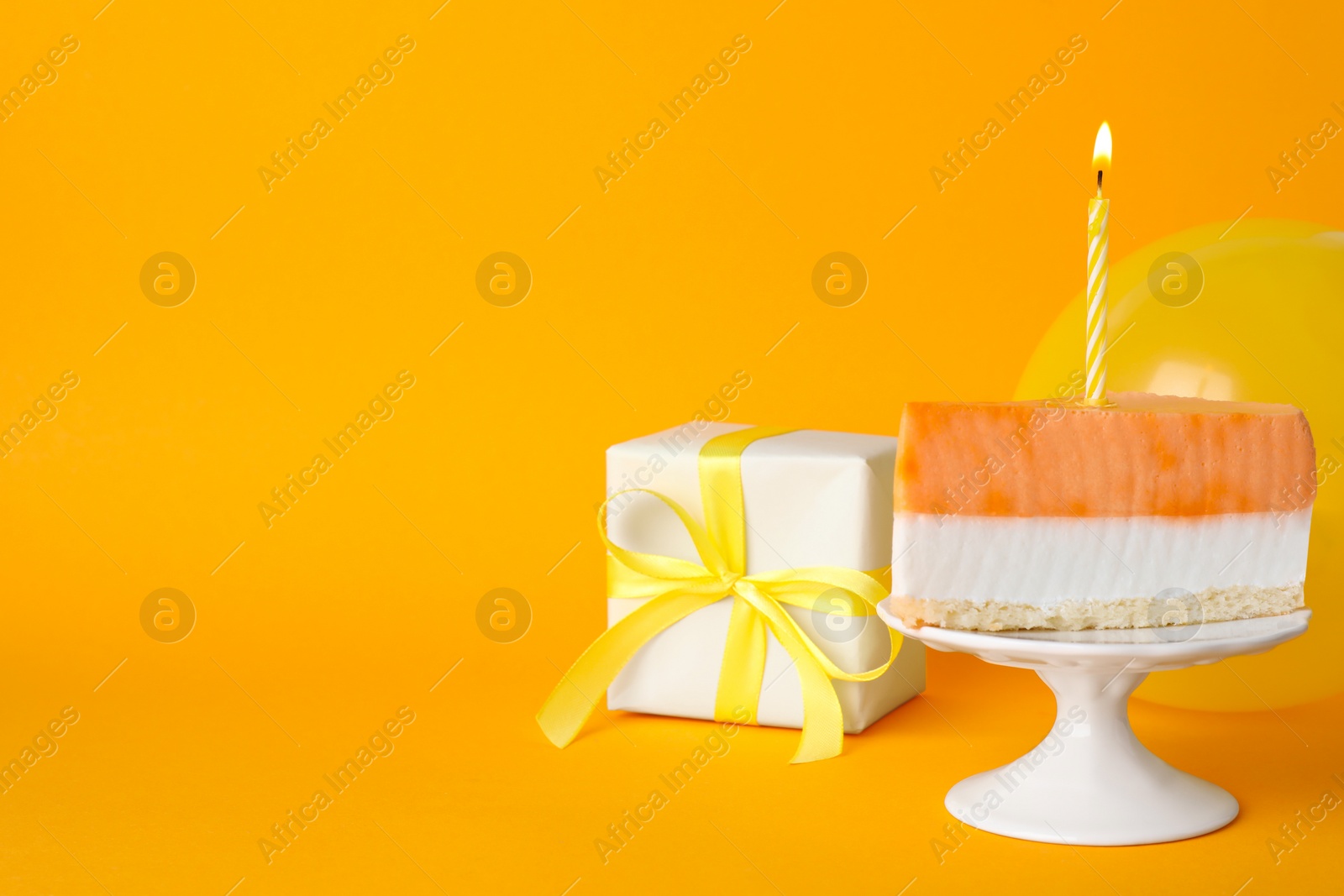 Photo of Piece of delicious birthday cake with candle near gift box and balloon on orange background, space for text