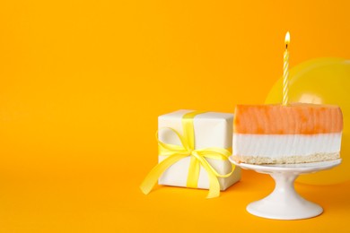 Piece of delicious birthday cake with candle near gift box and balloon on orange background, space for text