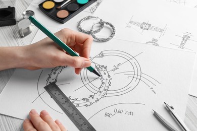 Photo of Jeweler drawing sketch of elegant earrings on paper at white wooden table, closeup