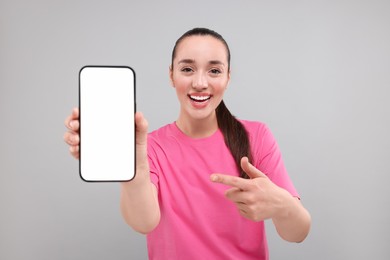 Photo of Young woman showing smartphone in hand and pointing at it on light grey background