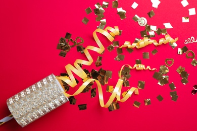 Golden confetti and streamers with party cracker on red background, top view