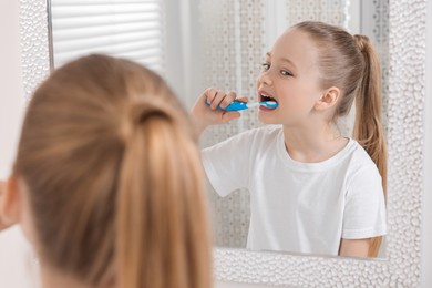 Photo of Cute little girl brushing her teeth with plastic toothbrush near mirror in bathroom