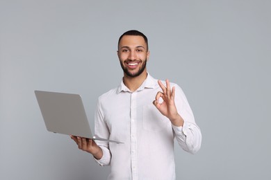 Photo of Smiling young man with laptop showing OK gesture on grey background