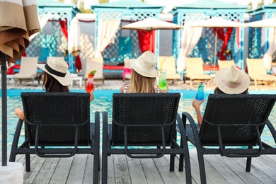 Photo of Happy young friends with refreshing cocktails relaxing on deck chairs near swimming pool