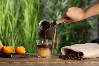 Photo of Woman pouring coffee into glass with orange juice and ice cubes at wooden table, closeup