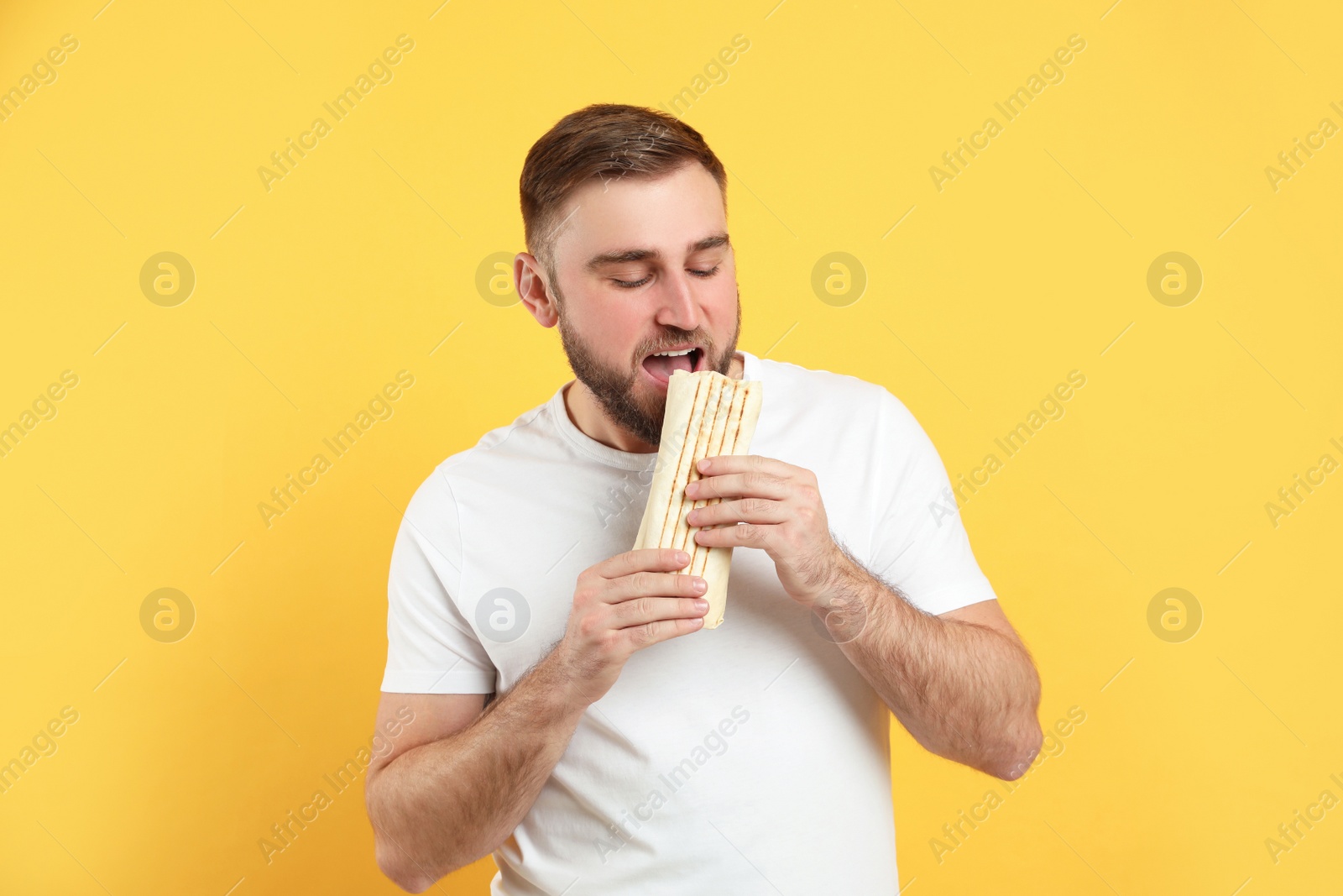 Photo of Young man eating delicious shawarma on yellow background