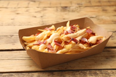 Photo of Tasty potato fries, cheese sauce and bacon in paper container on wooden table, closeup