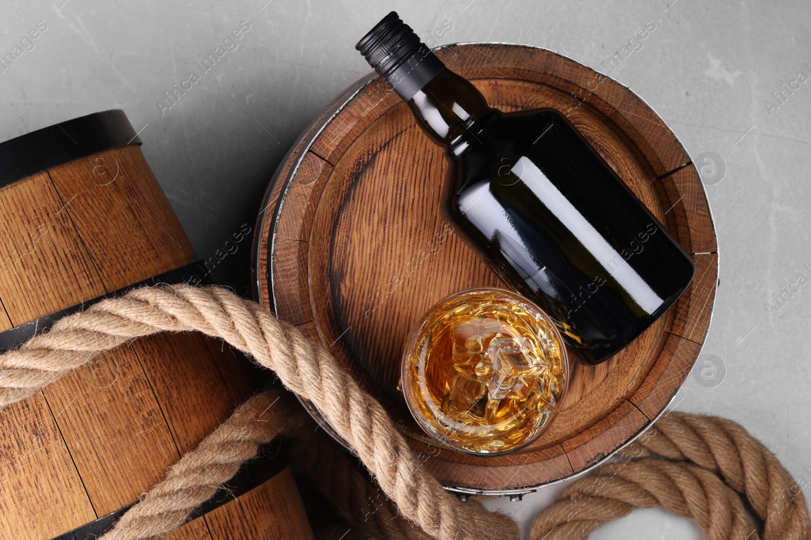 Photo of Whiskey with ice cubes in glass, bottle, wooden barrels and rope on grey marble table, top view