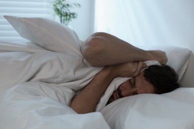 Photo of Sleepy man covering his ears with blanket in bed at home