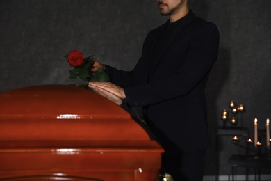Young man putting red rose onto casket lid in funeral home, closeup