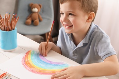 Photo of Cute little boy drawing rainbow with pencil at white wooden table in room. Child`s art