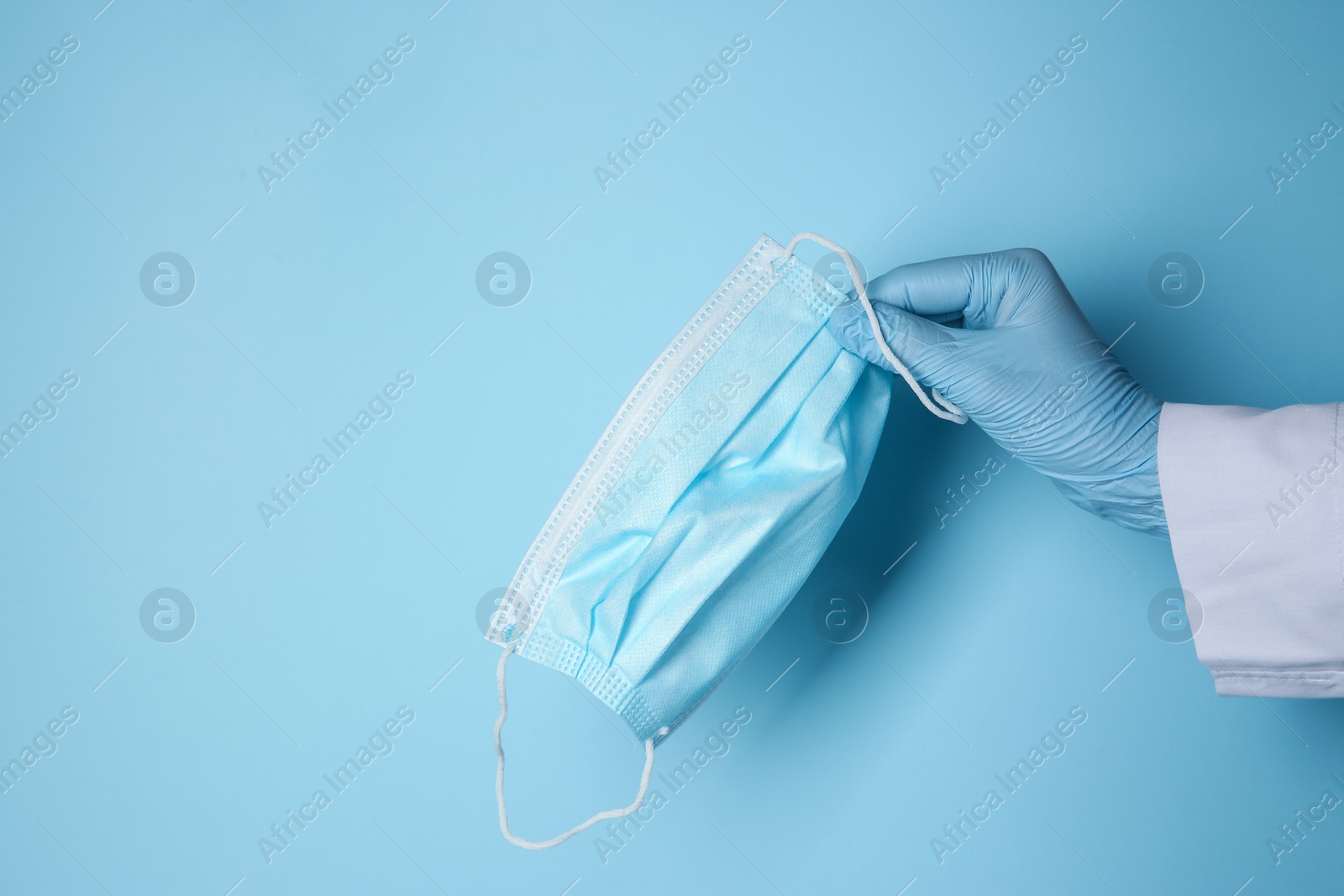 Photo of Doctor in latex gloves holding disposable face mask on light blue background, closeup with space for text. Protective measures during coronavirus quarantine