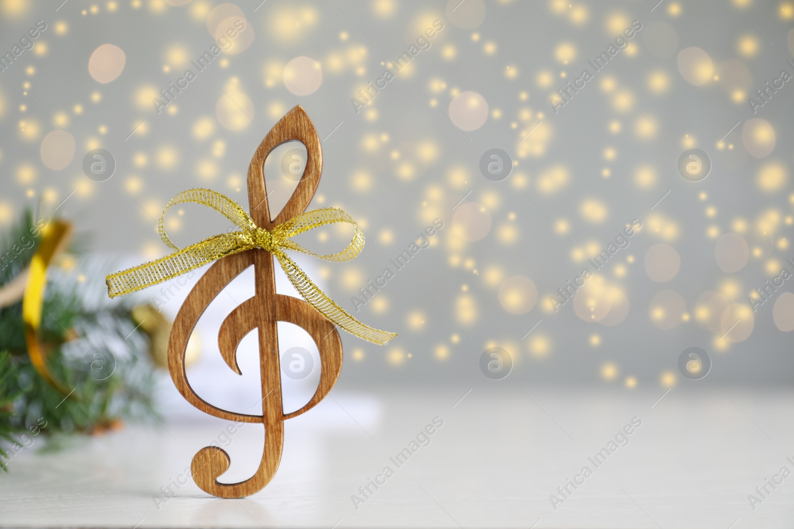 Photo of Wooden music note with golden bow on light grey table against blurred Christmas lights. Space for text