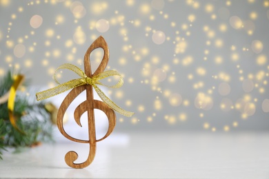 Photo of Wooden music note with golden bow on light grey table against blurred Christmas lights. Space for text