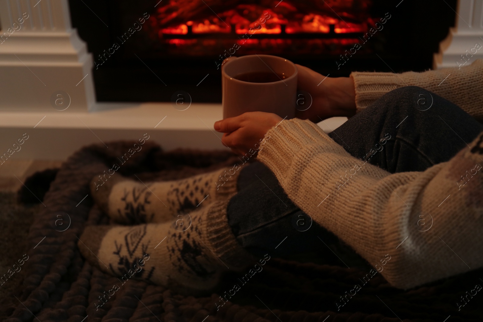 Photo of Woman in warm socks with cup of hot drink resting near fireplace at home, closeup