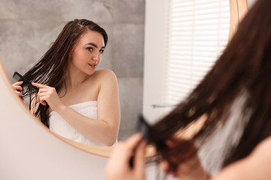 Photo of Young woman brushing hair after applying mask near mirror in bathroom