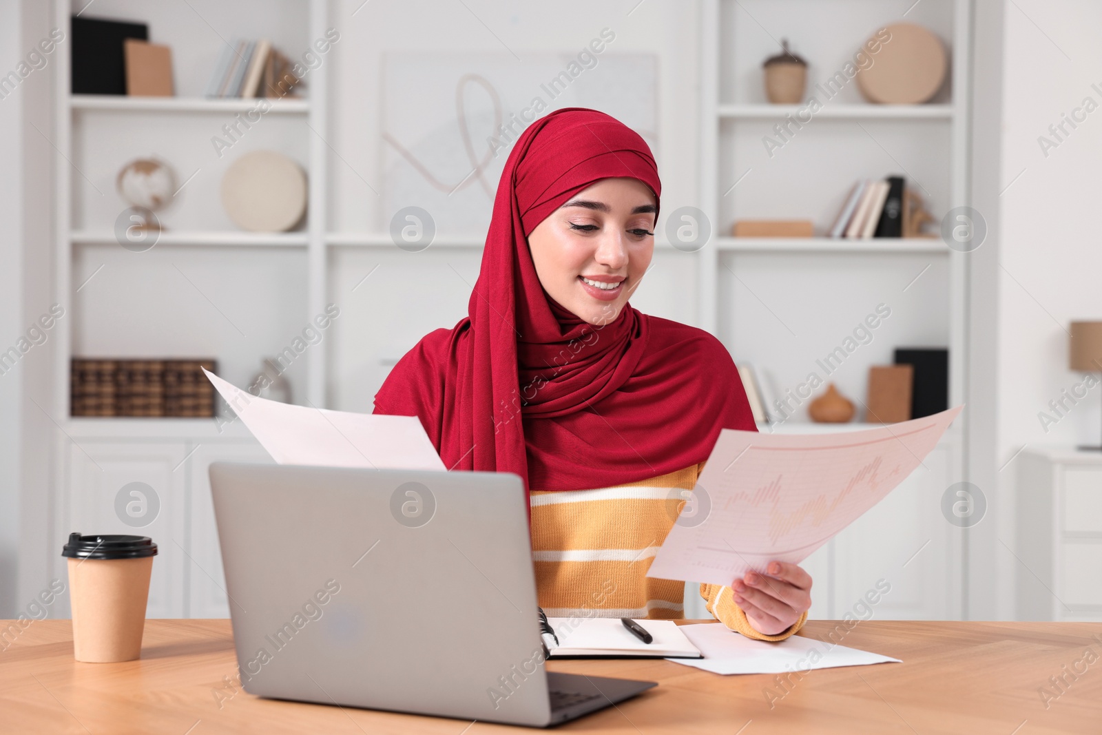 Photo of Muslim woman working near laptop at wooden table in room