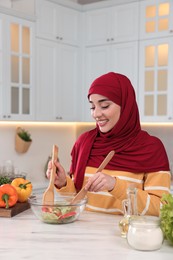 Photo of Muslim woman making delicious salad with vegetables at white table in kitchen