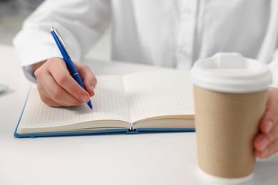 Young woman with cup of coffee writing in notebook at white table, closeup