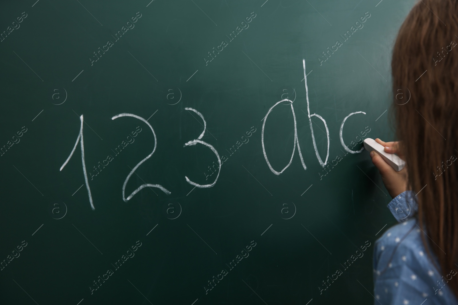 Photo of Little child writing letters and numbers on chalkboard, closeup