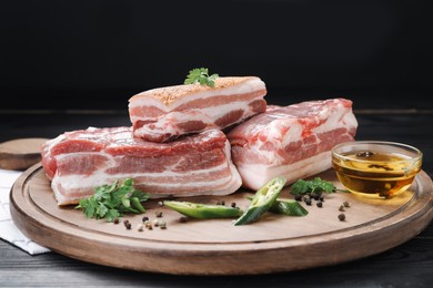 Photo of Pieces of raw pork belly, chili pepper, peppercorns, oil and parsley on black wooden table, closeup