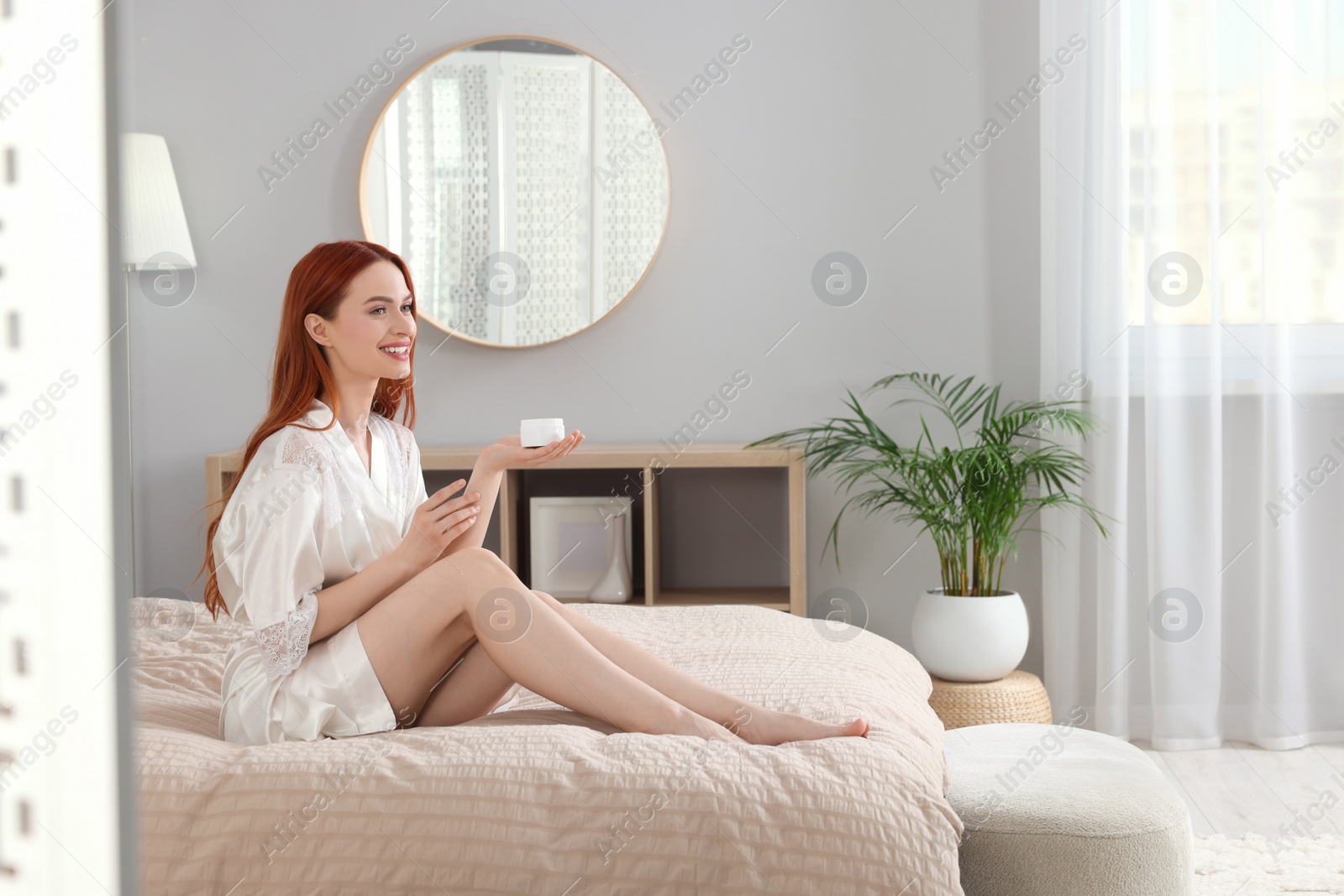 Photo of Beautiful young woman applying body cream onto arms in bedroom, space for text