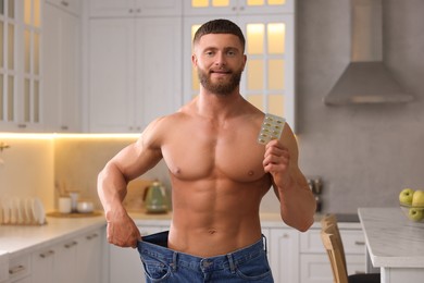 Photo of Athletic young man with pills wearing big jeans in kitchen. Weight loss