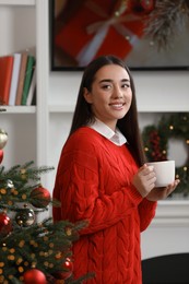 Smiling woman holding cup of hot drink near Christmas tree at home
