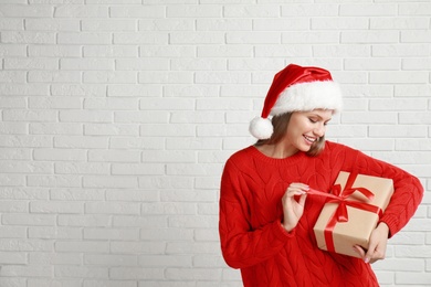 Happy young woman in Santa hat with Christmas gift near white brick wall. Space for text