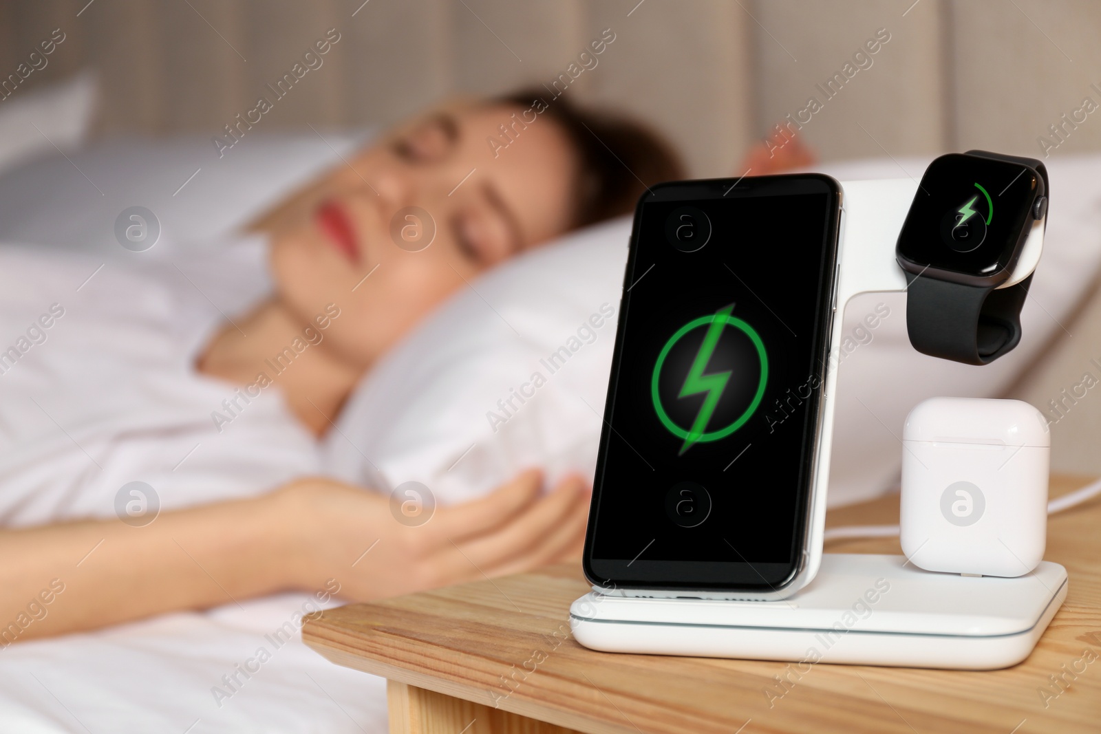 Photo of Smartphone, watch, earphones charging on wireless pad and sleeping woman in room