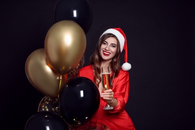 Photo of Happy woman in Santa hat with air balloons and glass of champagne on black background. Christmas party