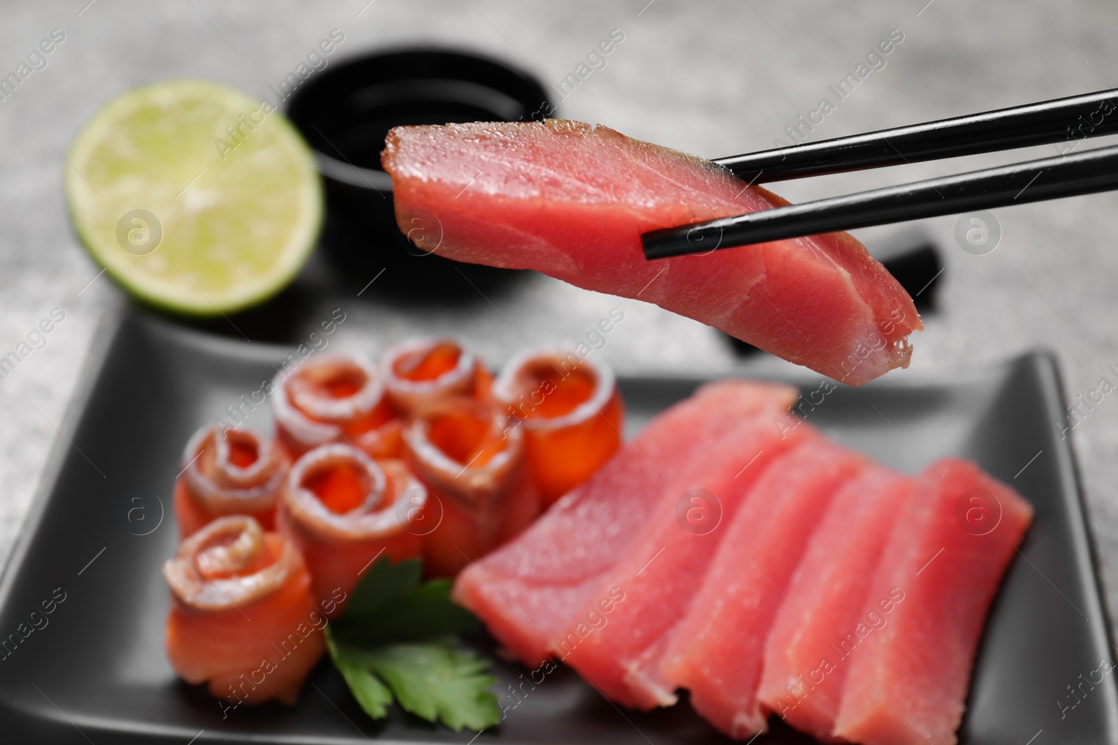 Photo of Chopsticks with raw tuna slice at grey table, closeup. Sashimi set served with soy sauce and lime