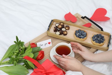 Photo of Tasty breakfast served in bed. Woman with tea, desserts, gift box, flowers and I Love You card at home, closeup