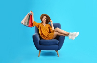 Photo of Beautiful young woman with paper shopping bags in armchair on light blue background