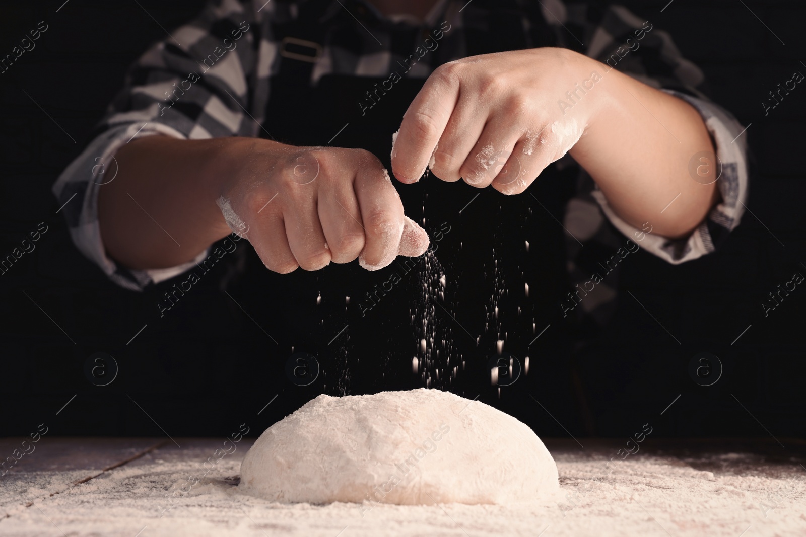 Photo of Man sprinkling flour over dough at wooden table on dark background, closeup