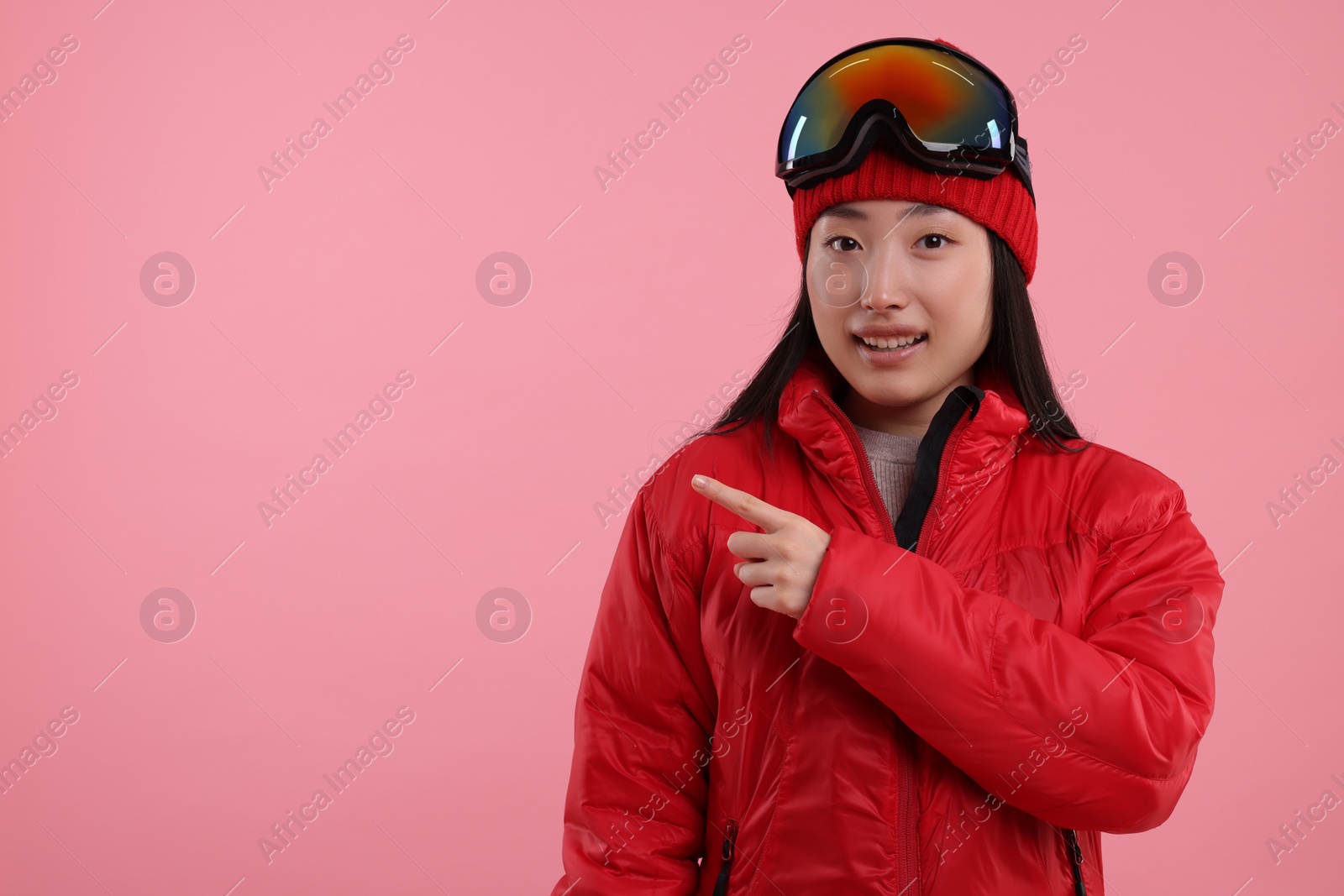 Photo of Happy woman in winter sportswear pointing at something on pink background. Space for text