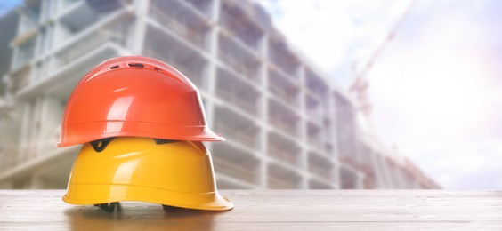 Image of Hard hats on wooden surface at construction site with unfinished building, banner design. Space for text 