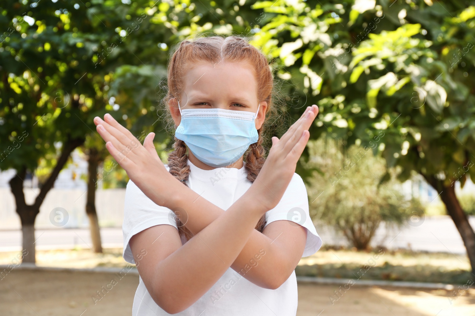 Photo of Little girl in protective mask showing stop gesture outdoors. Prevent spreading of coronavirus