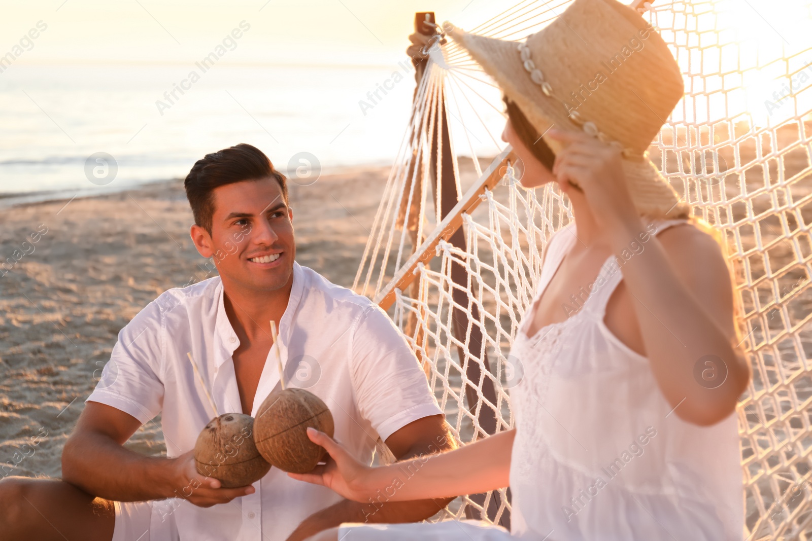 Photo of Young woman in hammock and her boyfriend with tropical cocktails outdoors at sunset