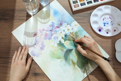 Photo of Woman painting flowers with watercolor at wooden table, above view. Creative artwork