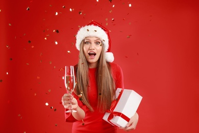 Emotional young woman in Santa hat with Christmas gift and glass of champagne on red background