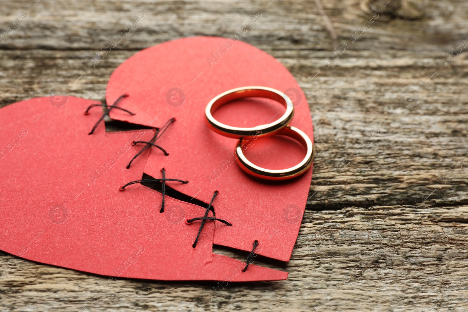 Photo of Broken heart. Torn red paper heart sewed with thread and wedding rings on wooden table, closeup. Space for text