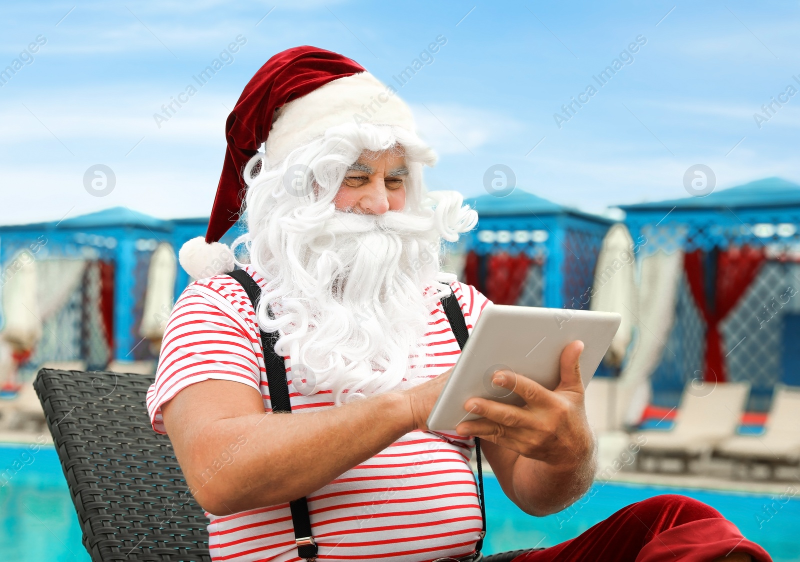 Photo of Authentic Santa Claus with tablet working on lounge chair at resort