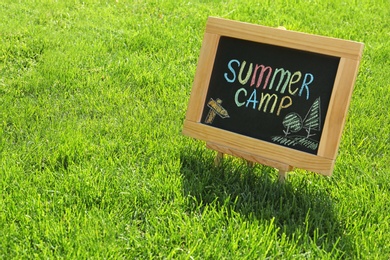 Photo of Blackboard with text SUMMER CAMP and drawings on green grass. Space for text