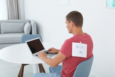 Man with note KICK ME on back working with laptop at table indoors. April fool's day prank