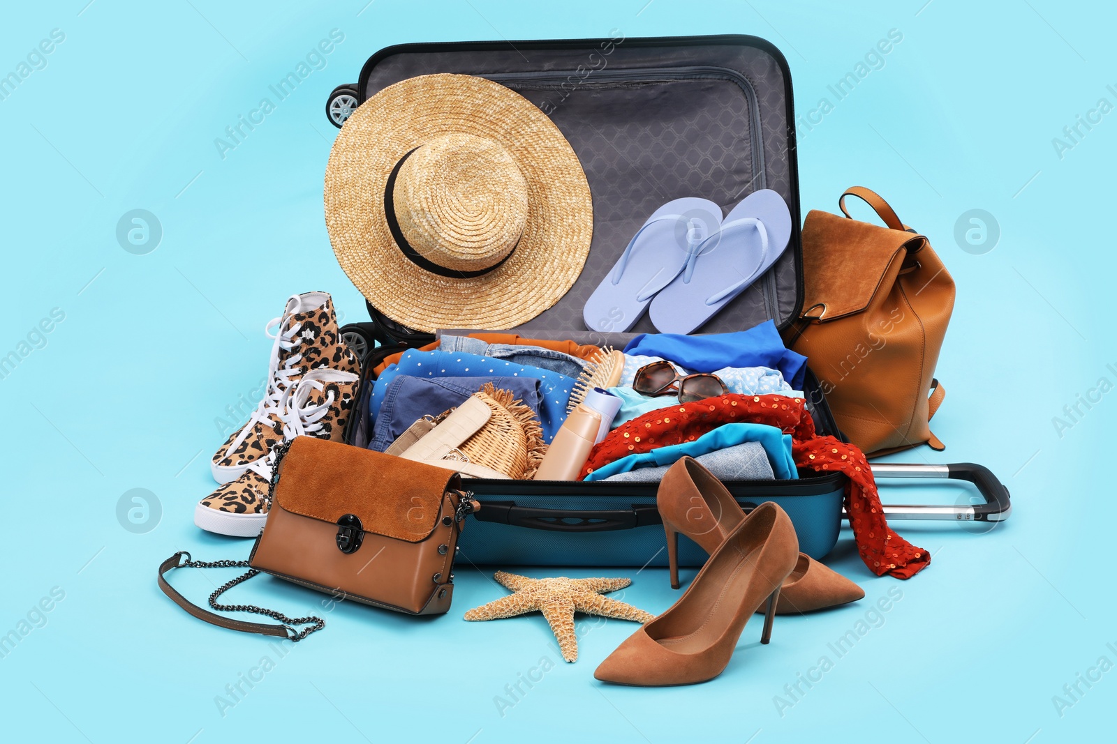 Photo of Open suitcase with clothes, beach accessories and shoes on light blue background. Summer vacation