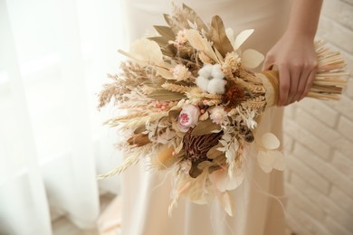 Bride holding beautiful dried flower bouquet near window at home, closeup