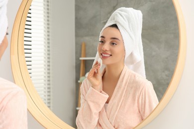 Photo of Beautiful woman in terry towel removing makeup with cotton pad near mirror indoors