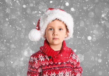 Cute child in Santa hat under snowfall on grey background. Christmas celebration