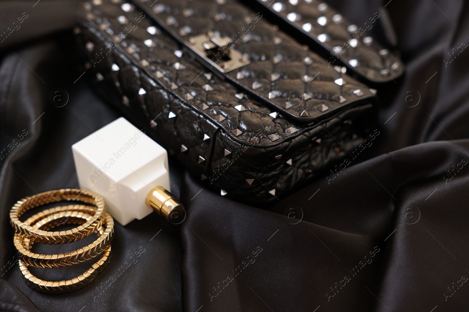 Photo of Leather bag, bottle of perfume and golden bracelets on black fabric, closeup